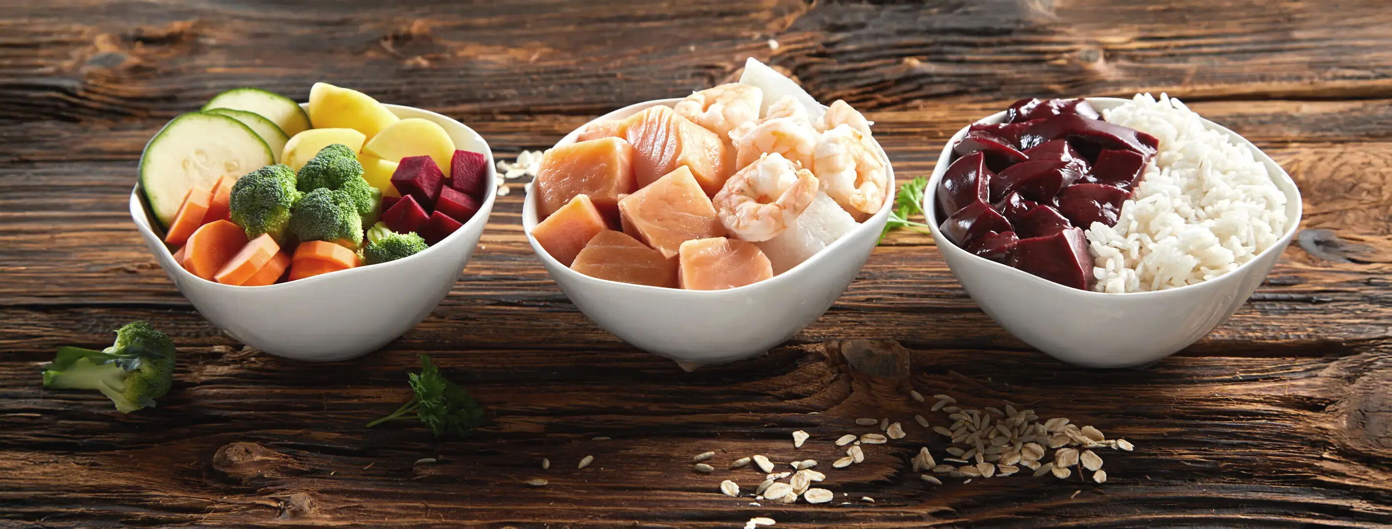 Drei Schüsseln auf einem Holztisch. Links eine Schüssel mit Gemüse: Zucchini, Brokkoli, Karotten, Rote Beete. In der Mitte eine Schüssel mit Lachs- und Garnelenstücken. Rechts eine Schüssel mit Reis und Rote-Bete-Stücken. Dazwischen liegen Haferflocken.