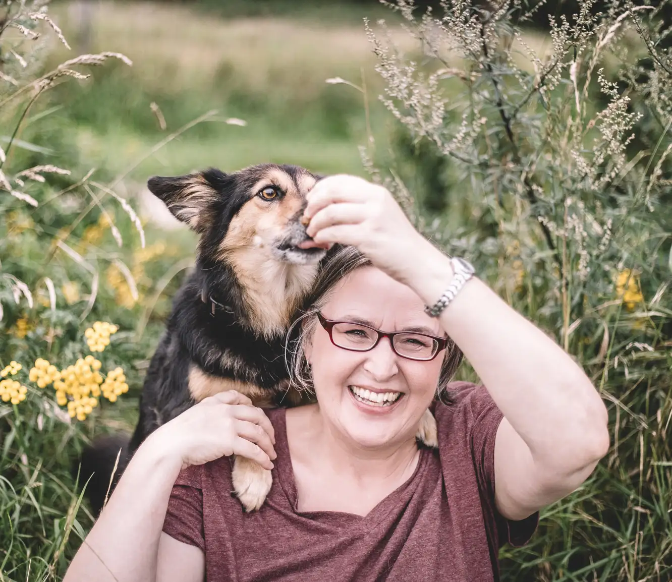Eine lachende Frau mit Brille sitzt in einer von Blumen und Gras umgebenen Wiese. Ein Corgi legt seine Pfoten auf ihre Schultern und schaut auf ihre Hand, die etwas hält. Die Szene ist entspannt und fröhlich.