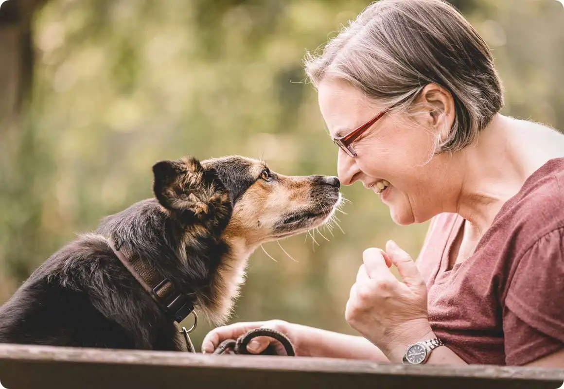 Eine ältere Person mit Brille lächelt und neigt sich nahe zu einem schwarz-braunen Hund, der ebenfalls interessiert blickt. Beide befinden sich draußen und haben eine freundliche Interaktion. Im Hintergrund ist unscharfes Grün zu sehen.